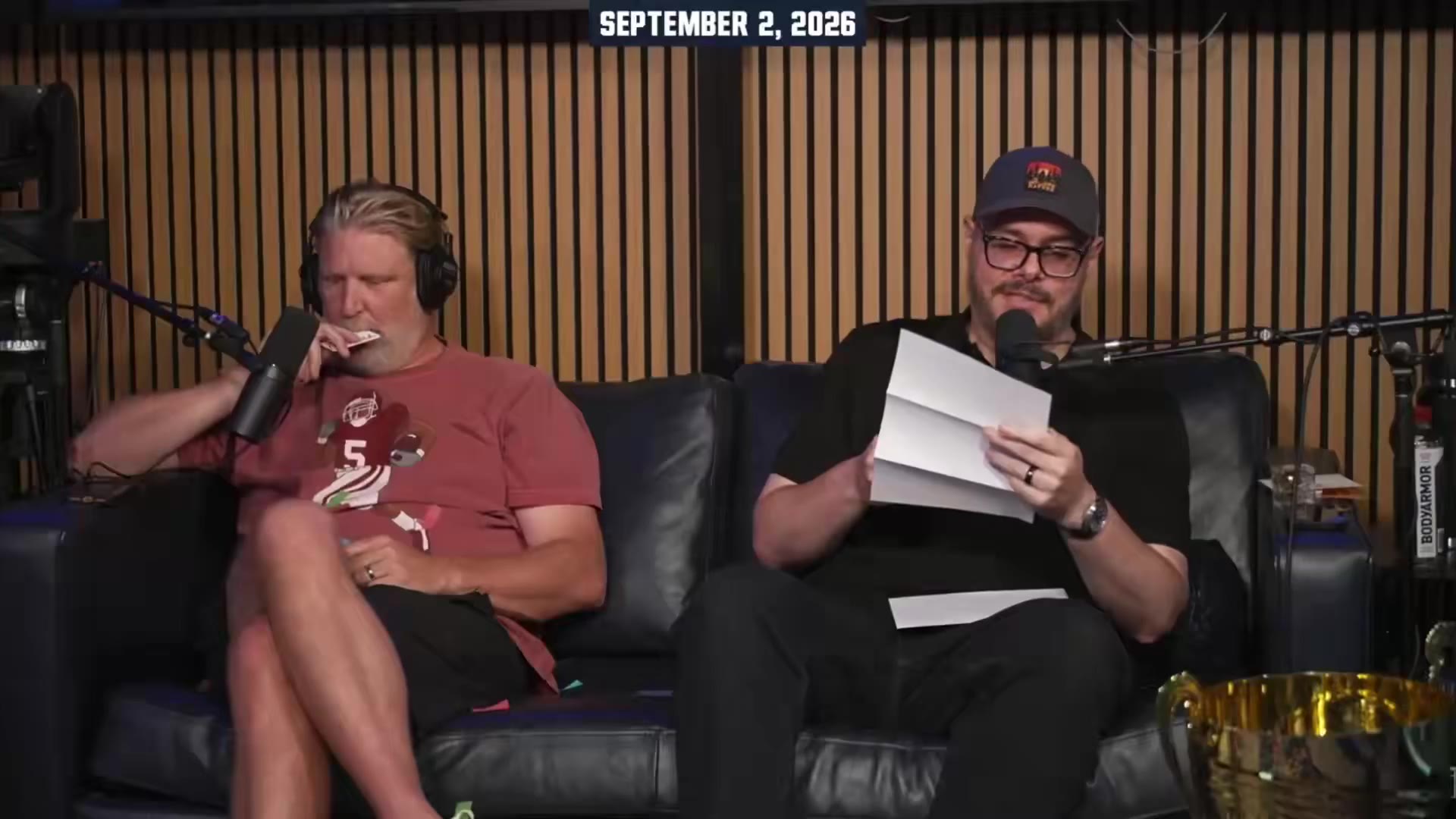 Two men sit on a leather couch in a podcast studio, one in a maroon t-shirt and shorts, the other in a black t-shirt, black cap, and glasses; a large gold trophy sits on the table between them, and confetti covers the man in maroon.