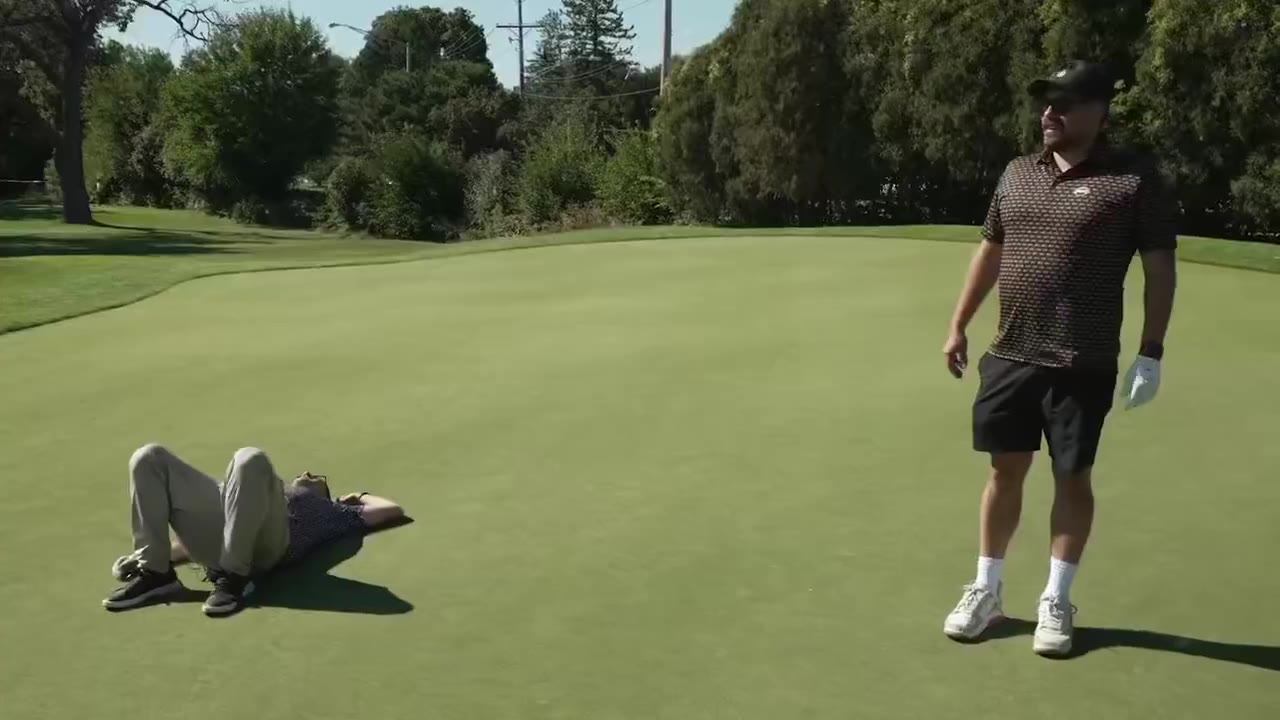 A man lies face-down on a golf green while another man in a striped polo and cap gestures nearby; a following shot shows a man driving a golf cart across the putting green with another cart visible in the background.