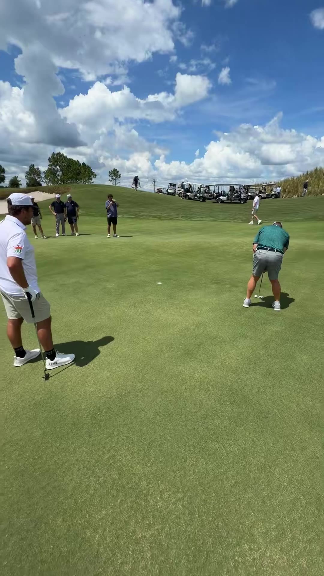 Two golfers stand on a putting green after finishing a hole, with other golfers, spectators, and golf carts visible in the background under a partly cloudy sky.