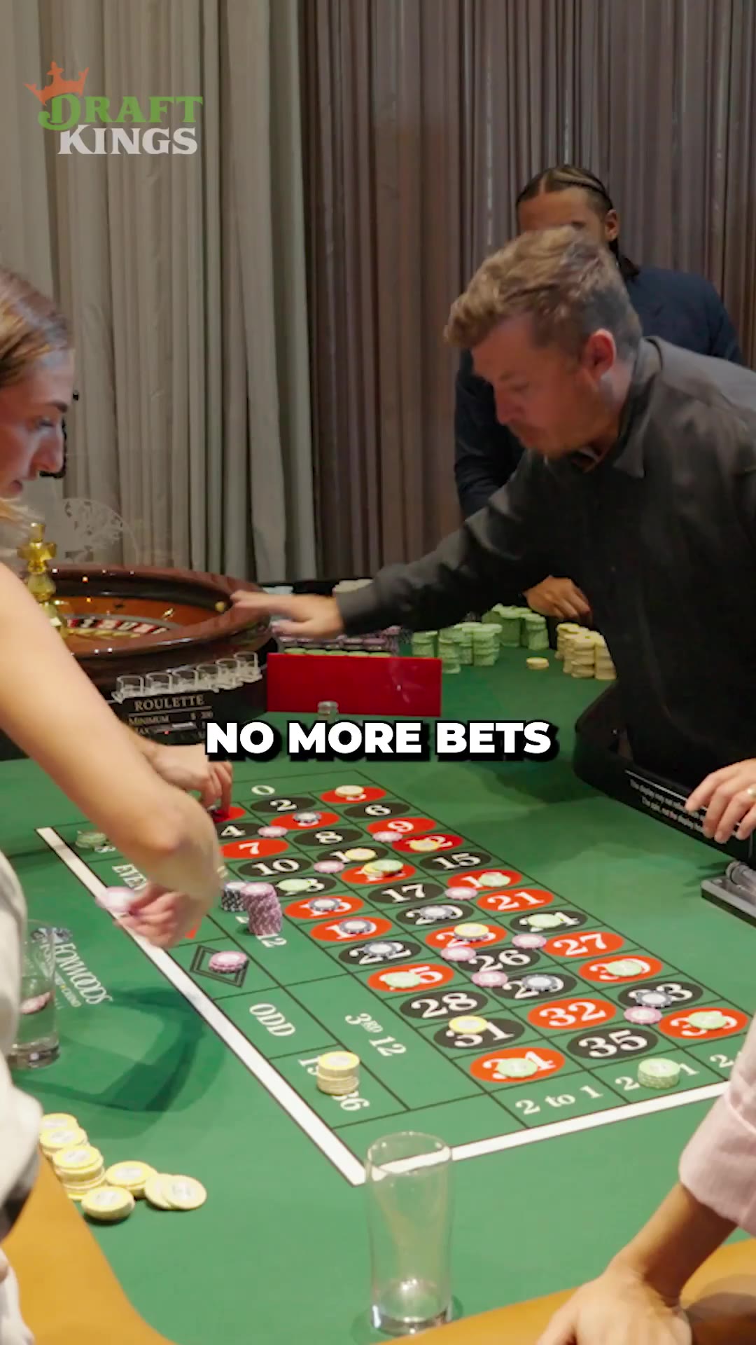 A group of men at a roulette table in a casino setting; on-screen text reads BLACK EVEN 22 as a man in a gray shirt places or handles chips on the layout while another man in a suit stands nearby, with a DraftKings logo in the corner and a split-screen comparison to a photo of former NBA player Patrick Ewing in a New York Knicks jersey.