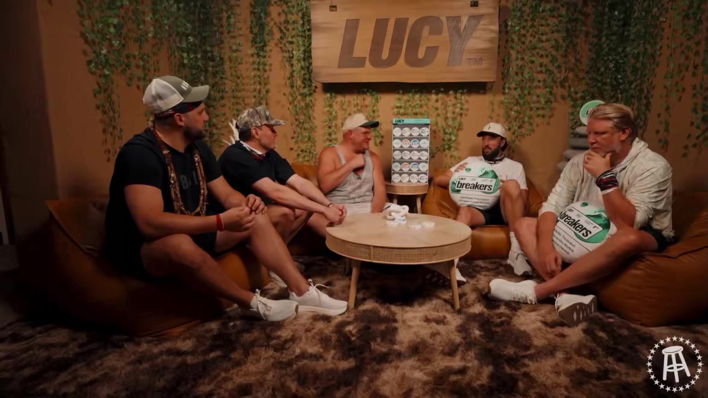 Six people sit in a lounge area near a fire pit, two wearing matching white "THE BOYS" t-shirts; one man in a black t-shirt and cap speaks while others listen; another man later holds up a large "LUCY breakers" nicotine pouch product display.