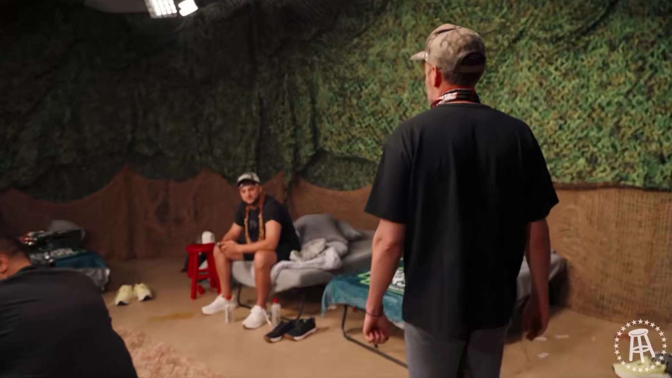 A group of people walk through a hotel lobby area with one saying 'Tommy: Do we have hotels tomorrow, or just tonight?', followed by two men sitting on camp cots in front of a camouflage netting backdrop, then a close-up of a man in a cap speaking in a dark interview setting.