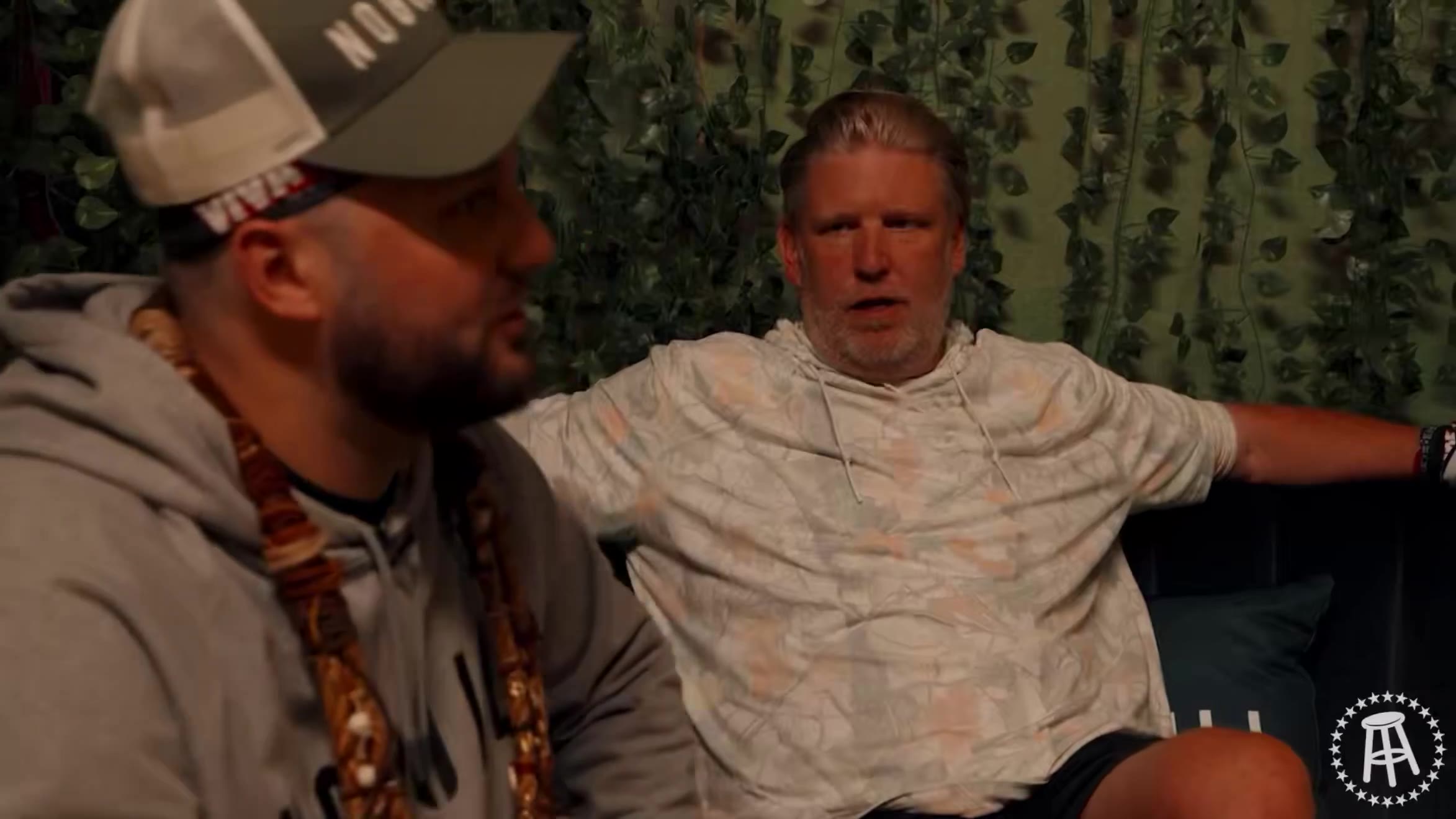Two men sit talking on a couch or bench in front of a green artificial-ivy wall, one wearing a trucker hat and hoodie with a beaded necklace, the other with gray hair wearing a tan patterned t-shirt and dark shorts, mid-conversation with subtitles on screen.