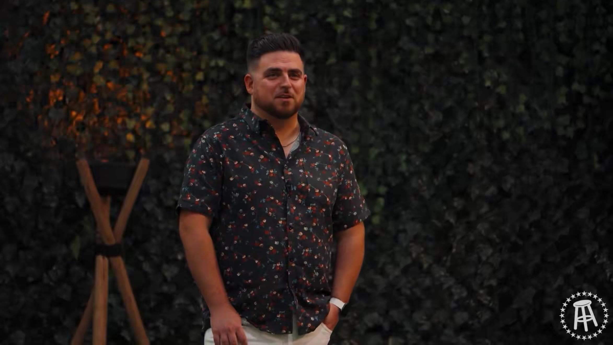 A bearded man in a white NOBULL t-shirt and cap stands on a Surviving Barstool set, followed by a shot of a man in a light patterned button-up shirt against a jungle-themed backdrop with wooden prop pillars.