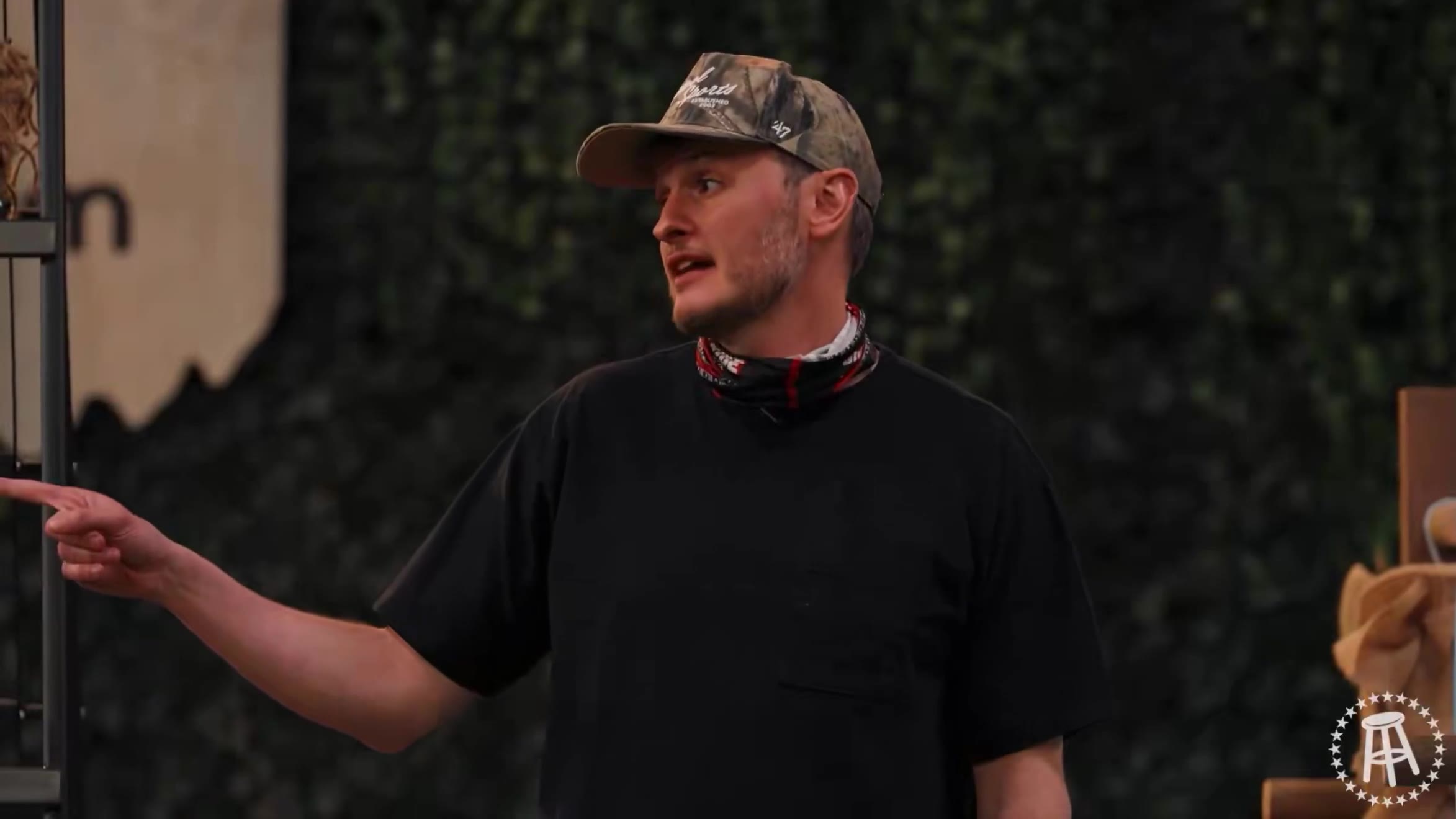 A man in a camo cap and black t-shirt speaks and gestures on an outdoor game-show style set with a rope ladder tower and a tetherball-style pole rig on green turf; a separate clip shows a man in a floral shirt holding what appears to be an animal tail or pelt in front of an ivy wall.