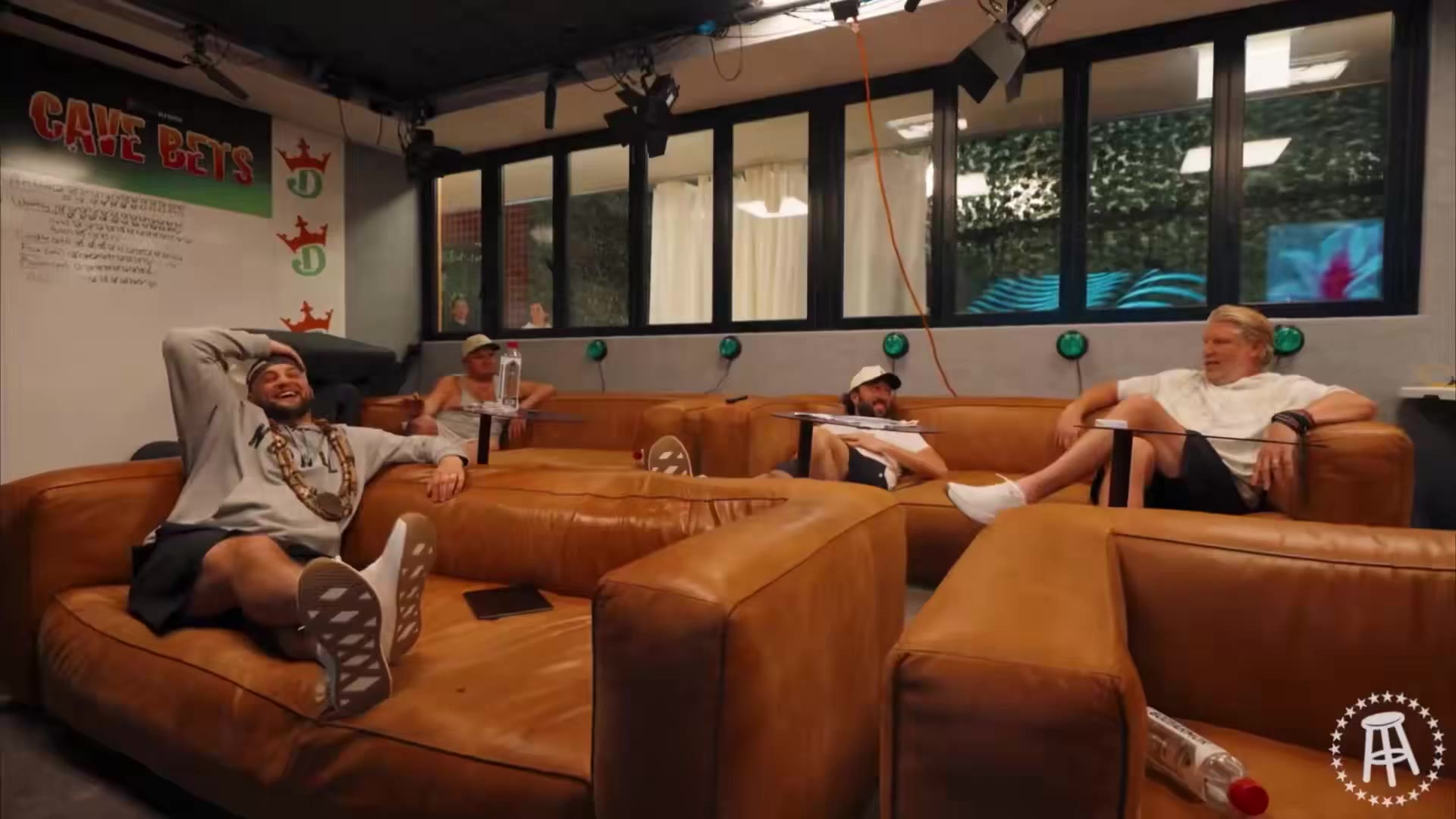 A group of men sit and talk on leather couches indoors, followed by a close-up of a blond man in a hoodie speaking to camera, followed by a man on an obstacle-course style set with a climbing net and padded discs.