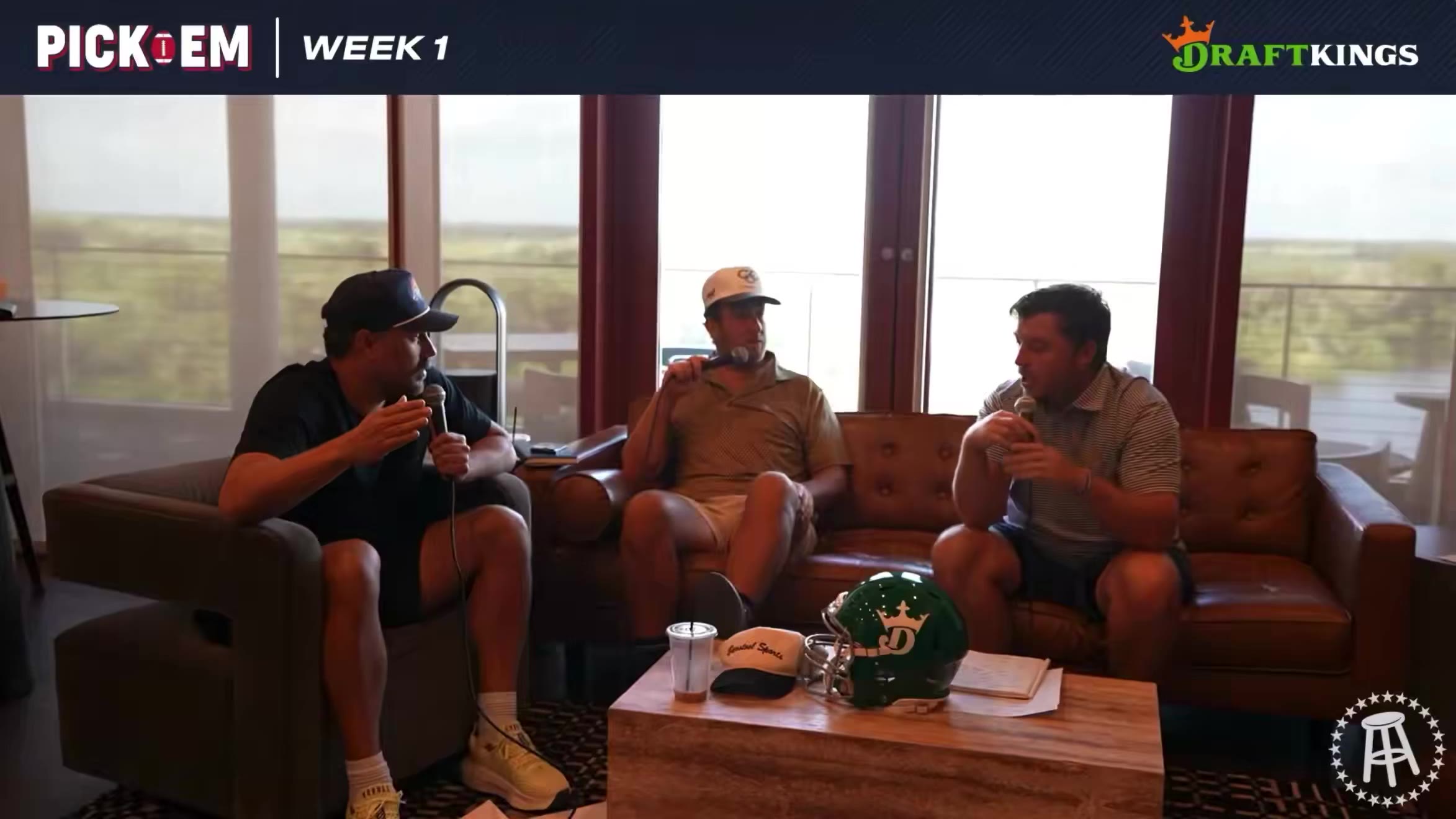 Two men sit on a leather couch in a room with large windows, each speaking into a wireless microphone; one wears a white cap and patterned polo, the other a striped polo, with a green football helmet resting on the floor between them and a DraftKings 'Pick 'Em Week 1' graphic banner across the top.