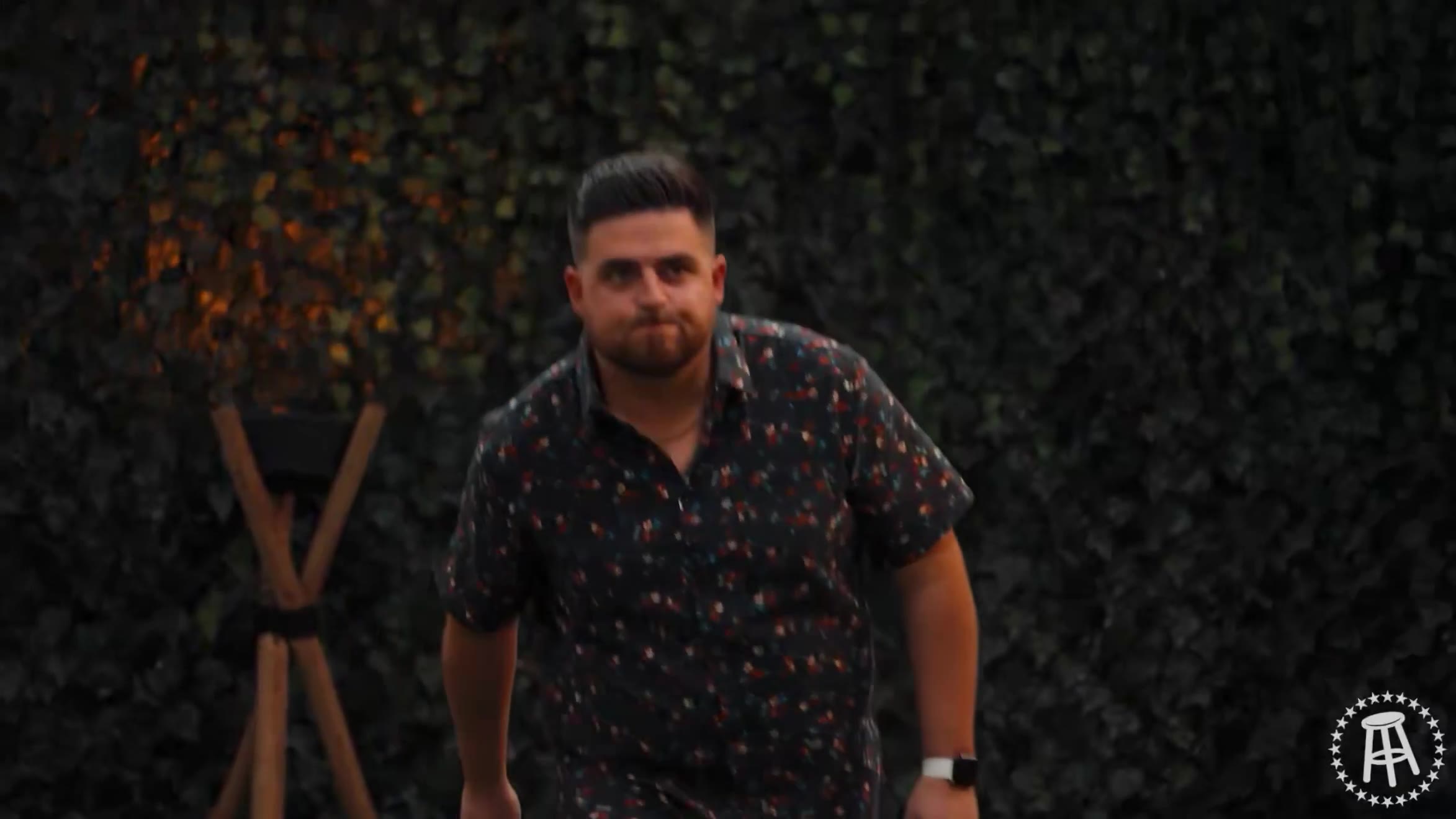 A contestant in a floral shirt walks across turf, followed by two men standing at obstacle-course stations labeled 'rumble premium' and 'Season 6 Surviving Barstool' with wooden platforms, poles, and yellow/blue prop pieces.