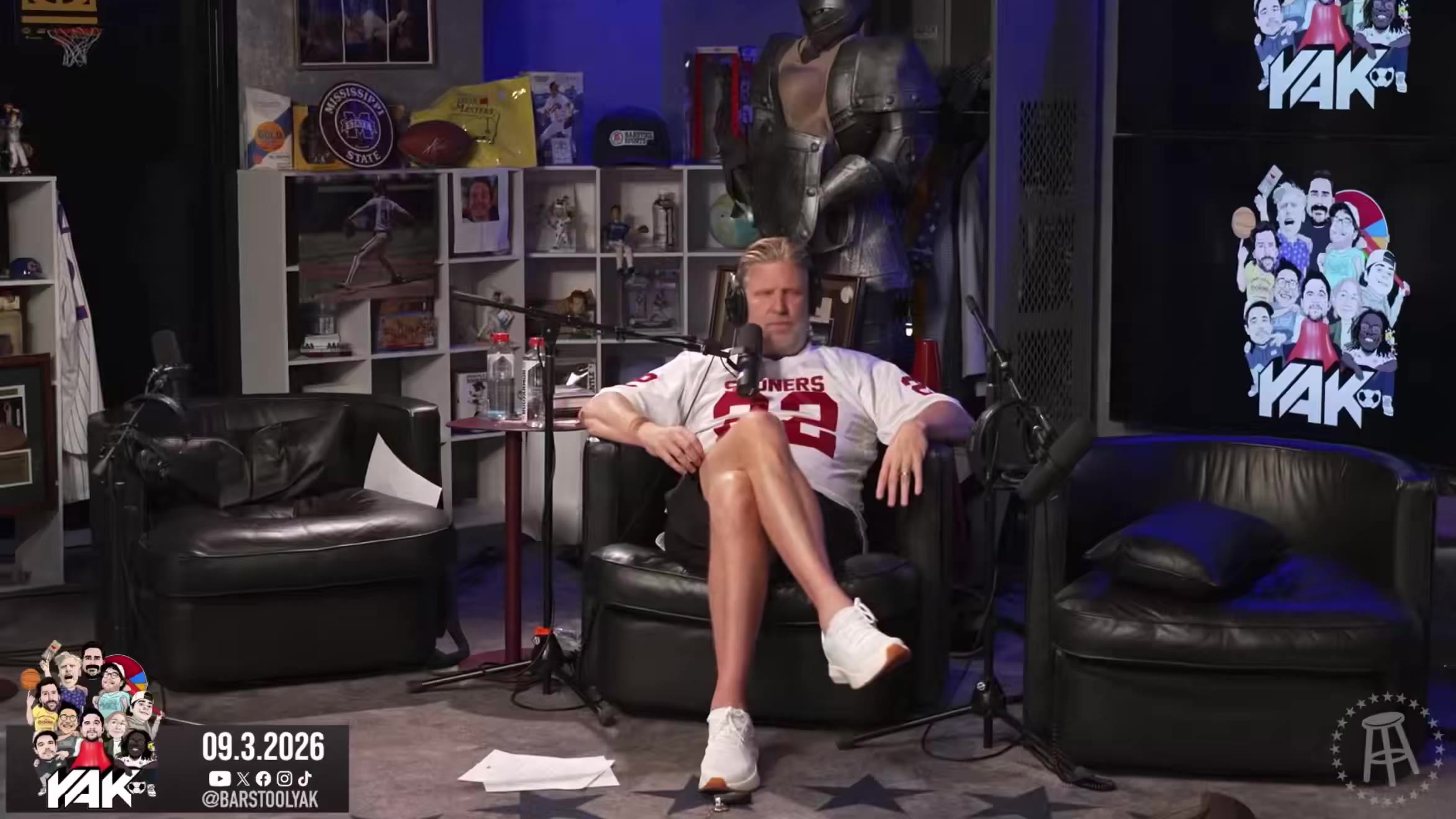 Two men in a podcast studio set with sports memorabilia and a suit of armor in the background; a seated man in a Sooners jersey talks into a microphone, then a standing man in a white 'ZAC'S ANGELS' crop top and black shorts gestures with his hands.