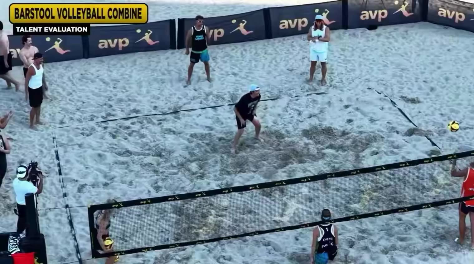 Multiple beach volleyball players in a sand court scrimmage under AVP-branded netting, with a bystander in a black t-shirt standing on the court holding a paddle-like object among the players; a later shot shows a group of women in black and purple outfits standing near the court sideline.