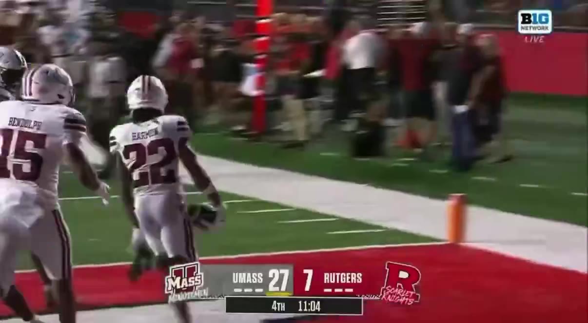 A UMass player runs into the end zone in front of celebrating white-jersey teammates on the sideline; a Rutgers staff member appears to fall or be knocked to the ground near the players; scoreboard reads UMass 27, Rutgers 7, 4th quarter 11:04.