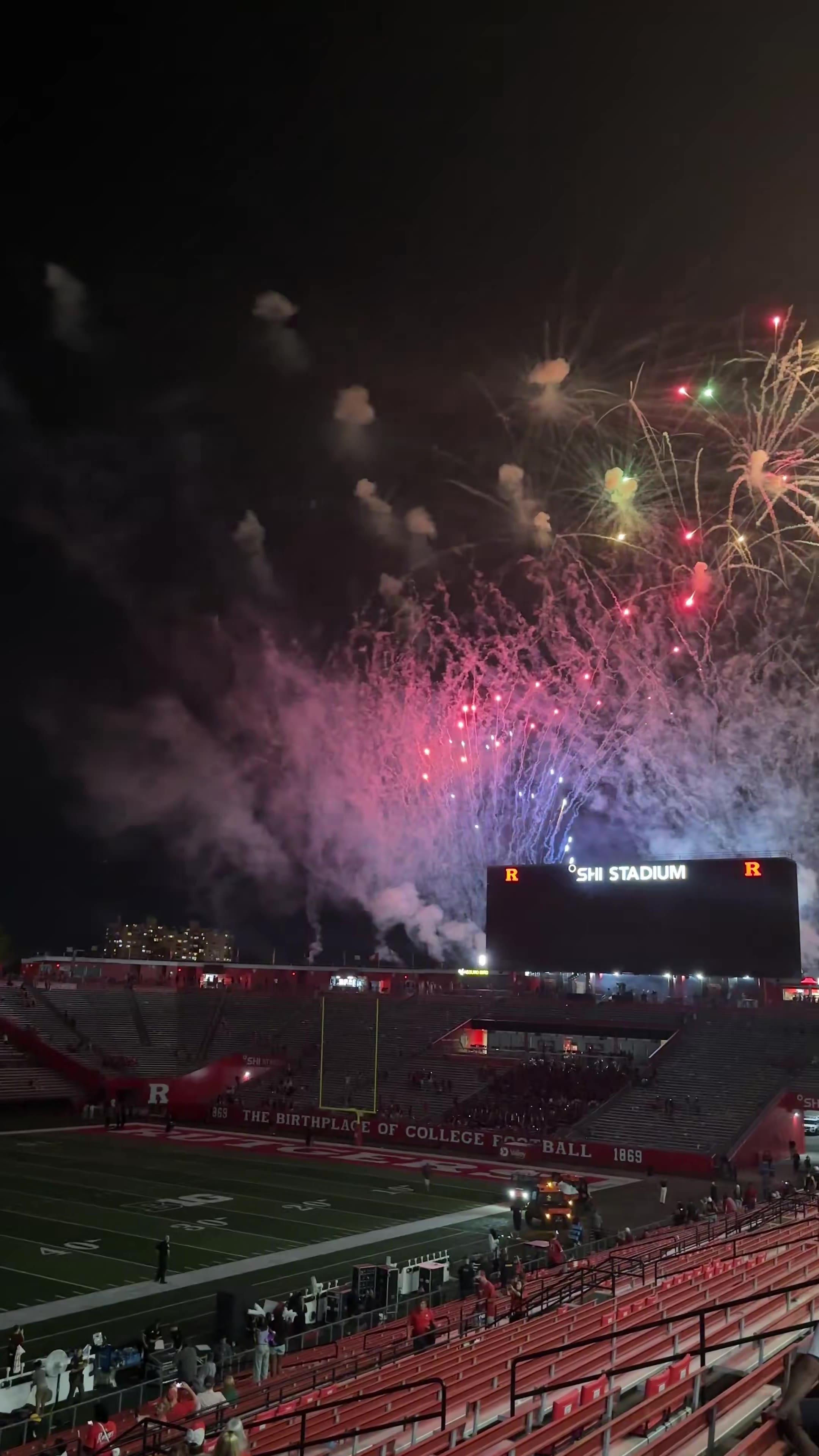 Fireworks explode over Rutgers' SHI Stadium at night as fans in the stands watch and film, with the stadium scoreboard reading SHI STADIUM visible below the empty upper bowl seating.