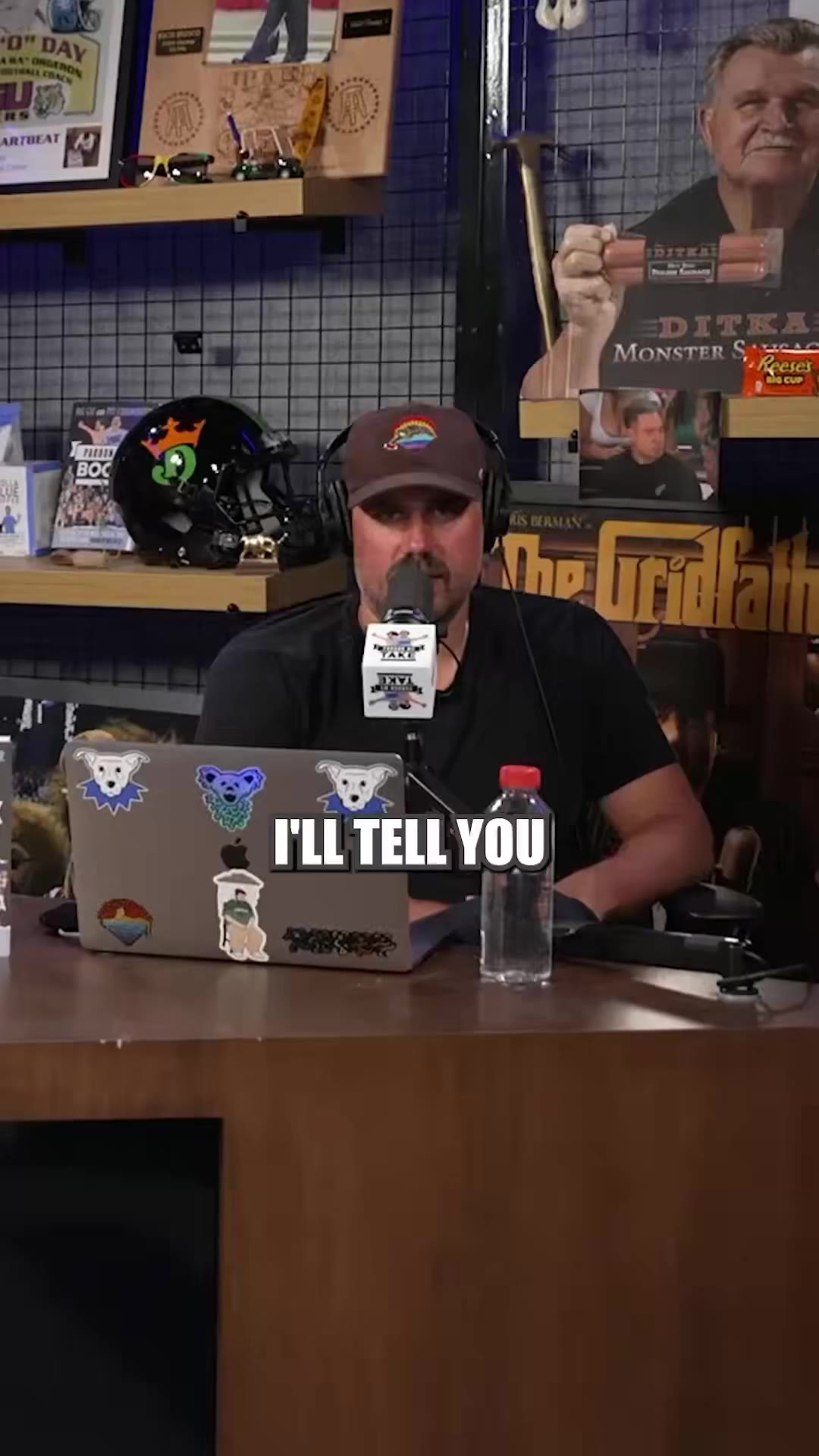 Two men wearing headphones sit at a Pardon My Take podcast desk in a room decorated with sports helmets and memorabilia, with on-screen captions reading STILL HURTS and CRAZY as one man speaks into a microphone and another laughs.
