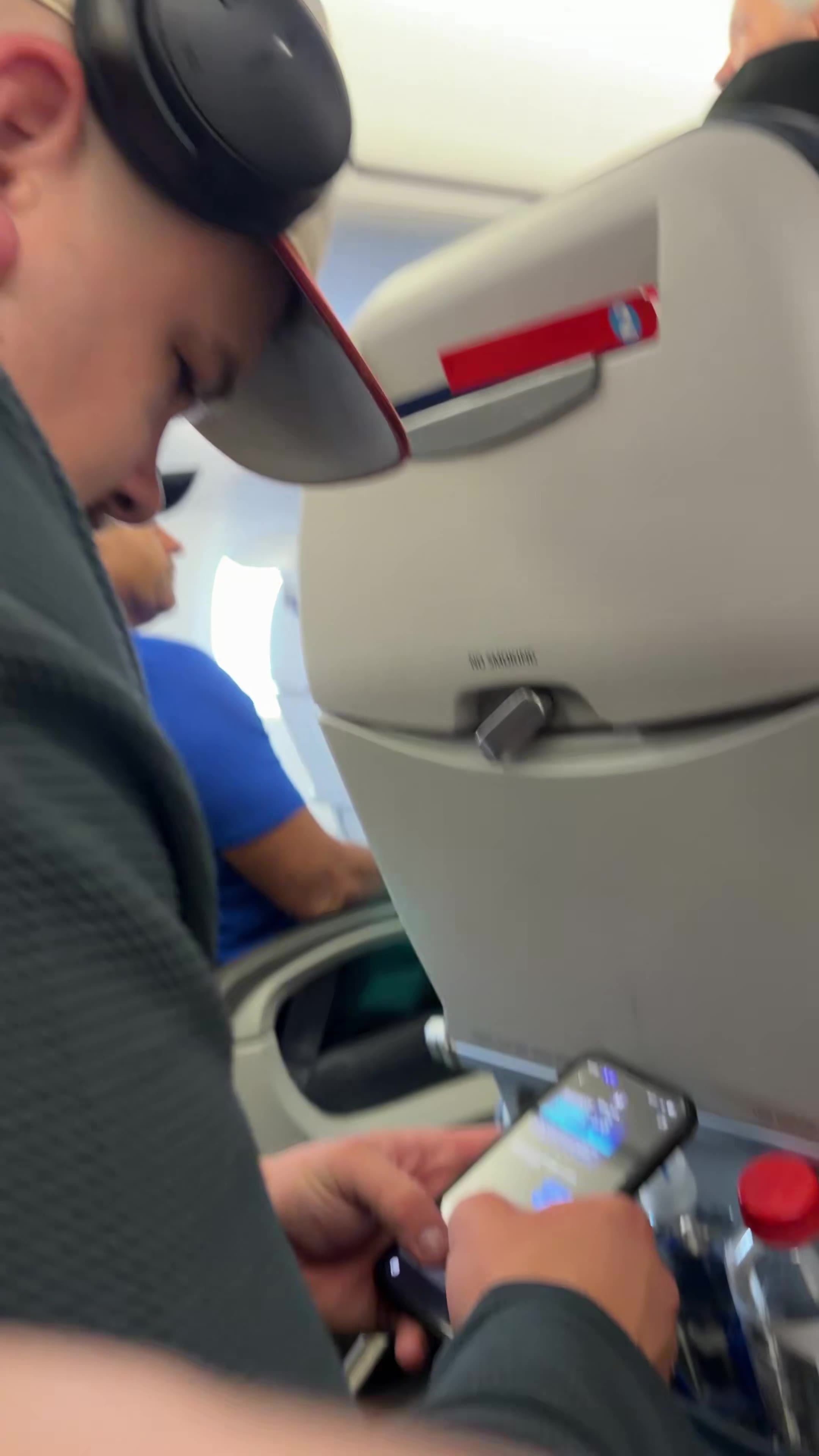 A person on a plane holds up a fist and points, then reaches up and touches their own cheek/jaw while wearing a cap and headphones, seated near another passenger in a blue shirt further up the aisle.