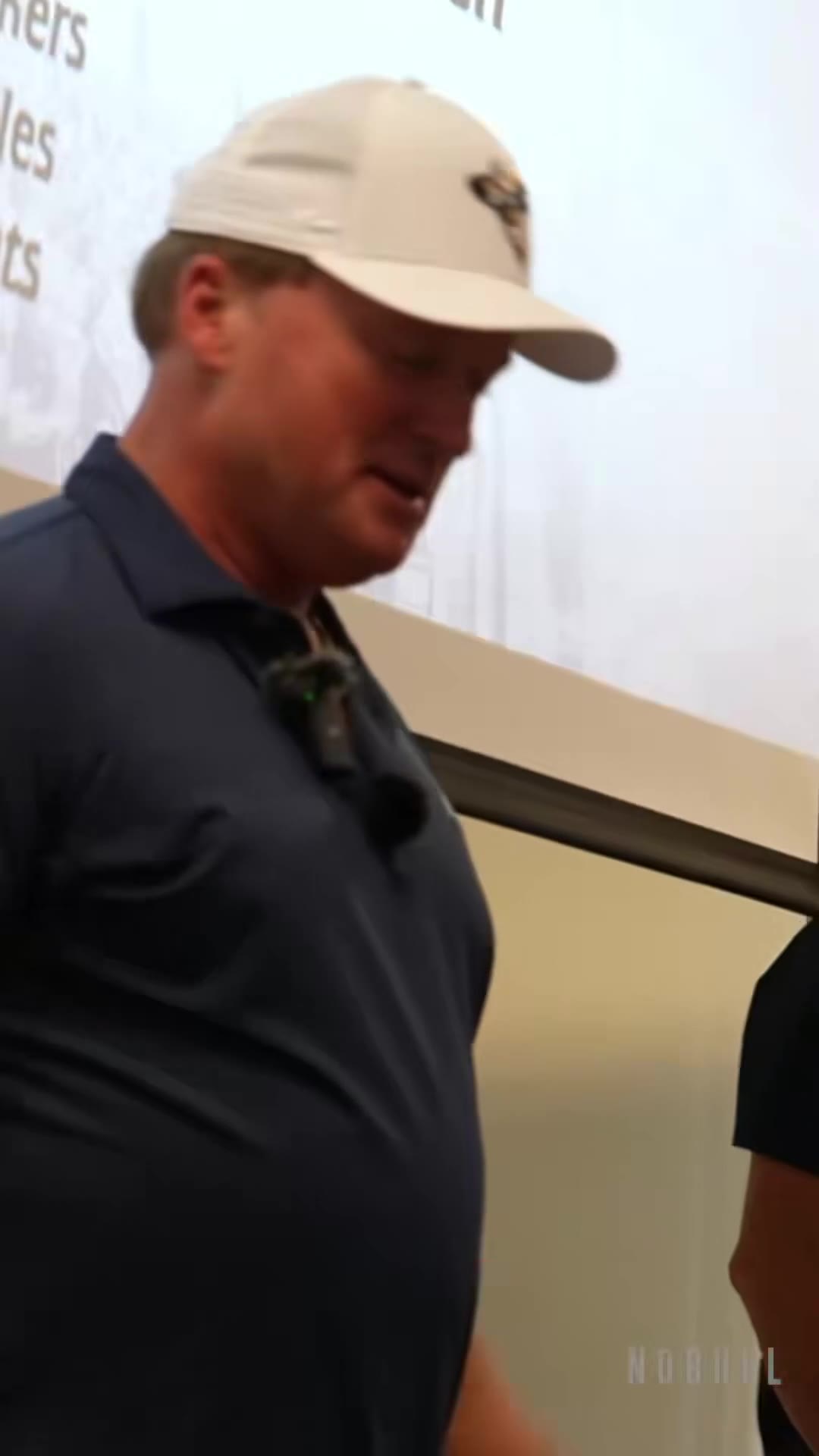 A man in a white cap and navy polo stands and speaks in an indoor room while others in matching navy polos stand nearby, near an illuminated red exit sign.