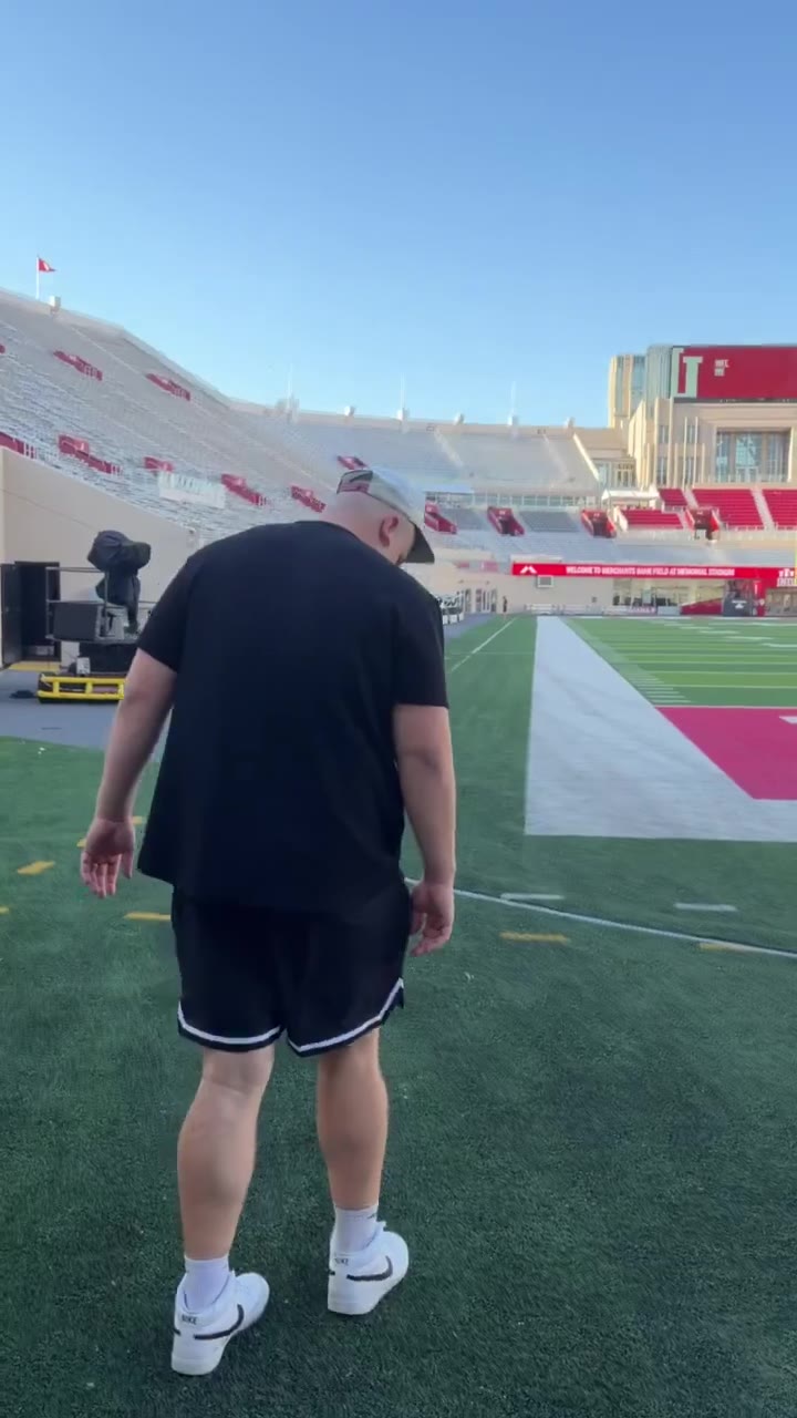 A man in a cap and black t-shirt walks along the sideline of an empty college football stadium at Indiana University's Memorial Stadium, with red bleacher seats and an 'Indiana University' sign visible on the upper deck.