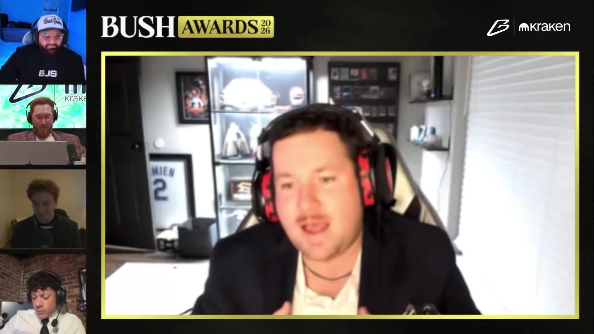 A man wearing headphones and a suit sits at a desk in a room with sports memorabilia and a display case, part of a multi-person video call graphic labeled 'BUSH AWARDS 2026'.