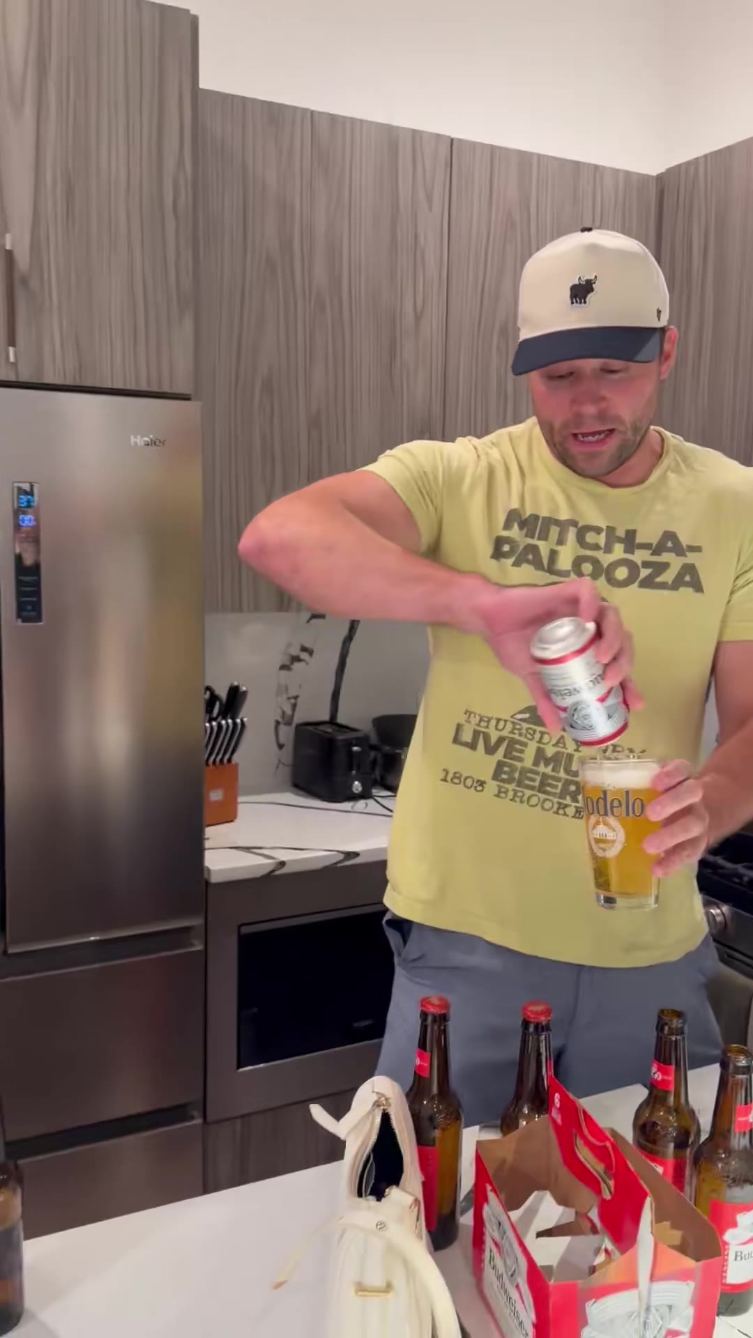 Two men in a kitchen shake hands and clink glasses over a case of beer, then a third younger man in a cap stands at a counter with his face buried in his hands.