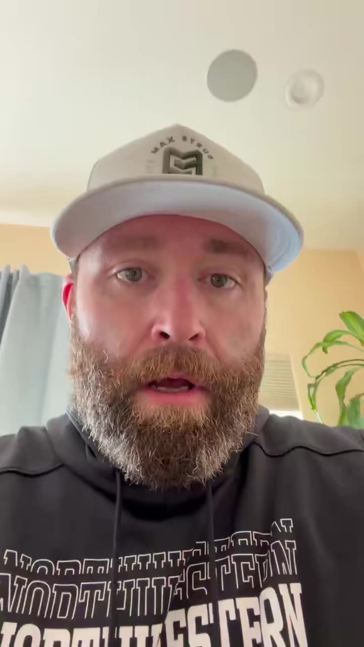A man wearing a white cap and a black Northwestern hoodie speaks directly to the camera in a bedroom or apartment setting, indoors with a curtain and plant visible in the background.